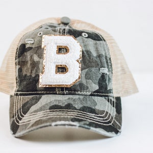 May include: A camouflage baseball cap with a white letter "B" embellished with gold sequins. The cap has a distressed look with a beige mesh back and a gray button on top. The brim is curved and the stitching is white.