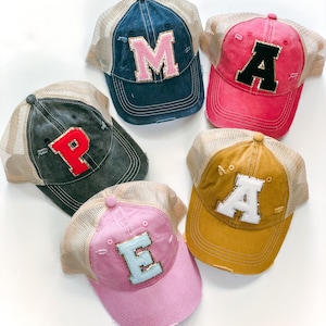 May include: Five distressed trucker hats in various colors, each featuring a different letter applique. Colors include navy, pink, mustard, and charcoal, with tan mesh backs. The letters are in contrasting colors, such as pink, black, and red.