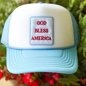 May include: A light blue and white trucker hat featuring a rectangular patch on the front panel. The patch has a light blue and white vertical striped background with the red embroidered text GOD BLESS AMERICA centered in three distinct lines.