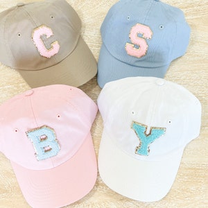 May include: Four baseball caps with different colored fabric and letters. The caps are beige, blue, pink, and white. The letters are C, S, B, and Y. The letters are made of a light blue material with gold glitter.