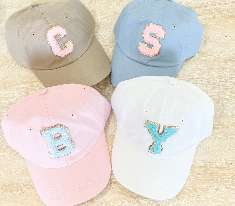 May include: Four baseball caps with different colored fabric and letters. The caps are beige, blue, pink, and white. The letters are C, S, B, and Y. The letters are made of a light blue material with gold glitter.