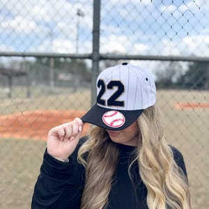 May include: Baseball cap with a black brim and white and black pinstripes. The cap features the number "22" in black with gold trim and a baseball patch. The background shows a baseball field and chain-link fence.