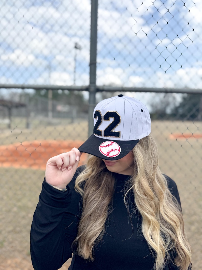May include: Baseball cap with a black brim and white and black pinstripes. The cap features the number "22" in black with gold trim and a baseball patch. The background shows a baseball field and chain-link fence.