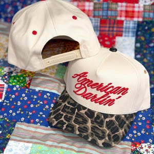 Puede incluir: Una gorra de b&eacute;isbol color crema con visera de estampado de leopardo y el texto bordado en rojo American Darlin' en la parte frontal. La gorra cuenta con ojales rojos en la corona y un cierre trasero de pl&aacute;stico ajustable tipo snapback.