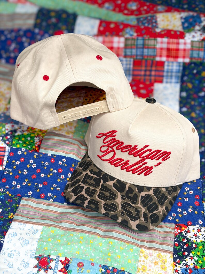Puede incluir: Una gorra de b&eacute;isbol color crema con visera de estampado de leopardo y el texto bordado en rojo American Darlin' en la parte frontal. La gorra cuenta con ojales rojos en la corona y un cierre trasero de pl&aacute;stico ajustable tipo snapback.