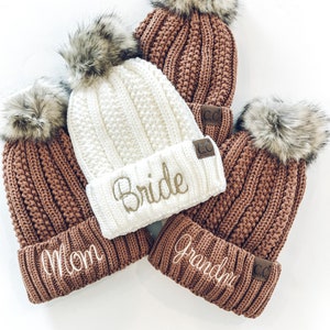 May include: Four knitted beanies with faux fur pom poms. The beanies are brown and white with embroidered text. The text reads "Mom", "Bride", "Grandma", and "CC".