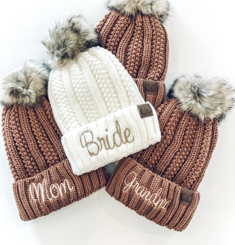 May include: Four knitted beanies with faux fur pom poms. The beanies are brown and white with embroidered text. The text reads "Mom", "Bride", "Grandma", and "CC".