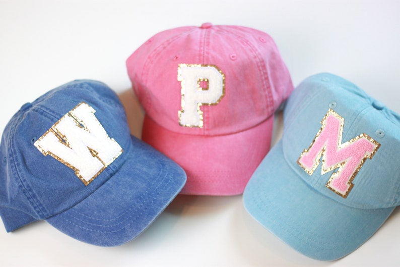 May include: Three baseball caps with different colored fabric and large white letters with gold glitter trim. The caps are blue, pink, and light blue. The letters on the caps are W, P, and M.