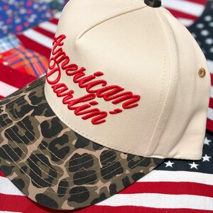 Puede incluir: Una gorra de b&eacute;isbol de color crema con visera de estampado de leopardo y el texto American Darlin' bordado en letra roja en la parte delantera. La gorra tiene un peque&ntilde;o bot&oacute;n oscuro en la parte superior y un ojal beige en el lateral.