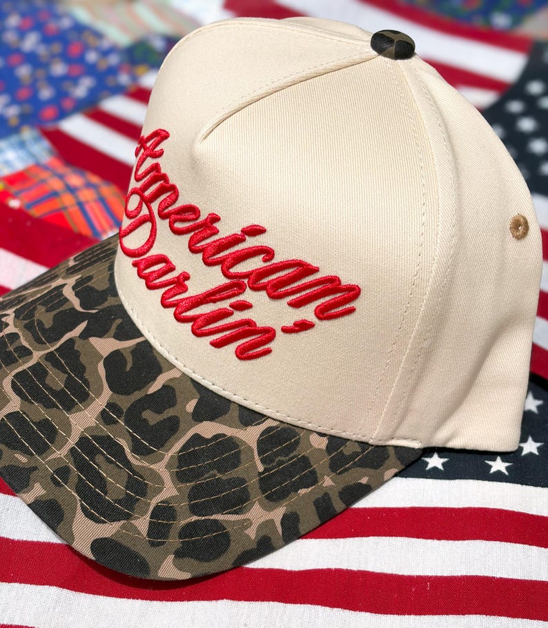 Puede incluir: Una gorra de b&eacute;isbol de color crema con visera de estampado de leopardo y el texto American Darlin' bordado en letra roja en la parte delantera. La gorra tiene un peque&ntilde;o bot&oacute;n oscuro en la parte superior y un ojal beige en el lateral.