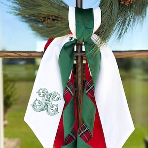 Puede incluir: Un lazo decorativo con paneles de tela blanca, uno de los cuales está bordado con una letra H ornamentada en verde. El lazo incluye cintas superpuestas en rojo sólido, verde oscuro sólido y un patrón de cuadros rojo, verde y azul.