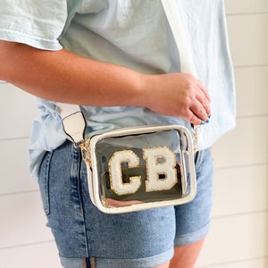 Puede incluir: Un bolso transparente de pl&aacute;stico con una bandolera ajustable blanca. El bolso tiene un monograma "CB" blanco y dorado.