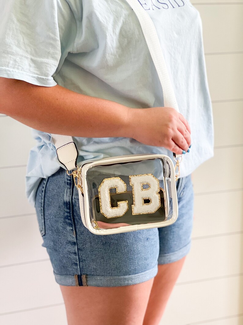 Puede incluir: Un bolso transparente de pl&aacute;stico con una bandolera ajustable blanca. El bolso tiene un monograma "CB" blanco y dorado.