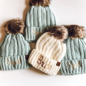 May include: Four knitted hats with faux fur pom poms. The hats are in shades of green and white. The hats are embroidered with the words "Squad" and "Bride".