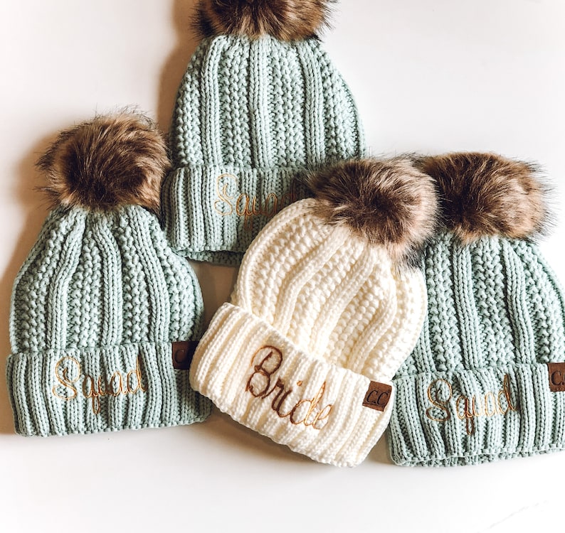 May include: Four knitted hats with faux fur pom poms. The hats are in shades of green and white. The hats are embroidered with the words "Squad" and "Bride".