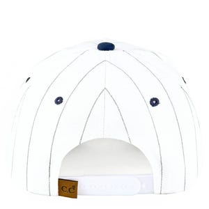 May include: White baseball cap with navy blue pinstripes and a navy blue button on top. The cap has a curved brim and a white adjustable snap closure. A brown leather tag with the letters "c.c" is attached to the back.