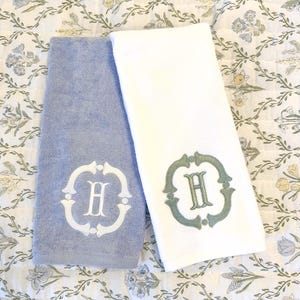 May include: Two plush hand towels, one light blue and one white, are displayed. Each towel features an embroidered letter "H" within a decorative frame. The towels are resting on a floral patterned fabric.