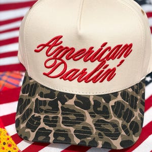 Puede incluir: Una gorra de b&eacute;isbol de color crema con el texto rojo bordado American Darlin' en el panel frontal. La visera de la gorra muestra un patr&oacute;n de estampado de leopardo marr&oacute;n y negro, y un peque&ntilde;o bot&oacute;n a juego se encuentra en la parte superior.