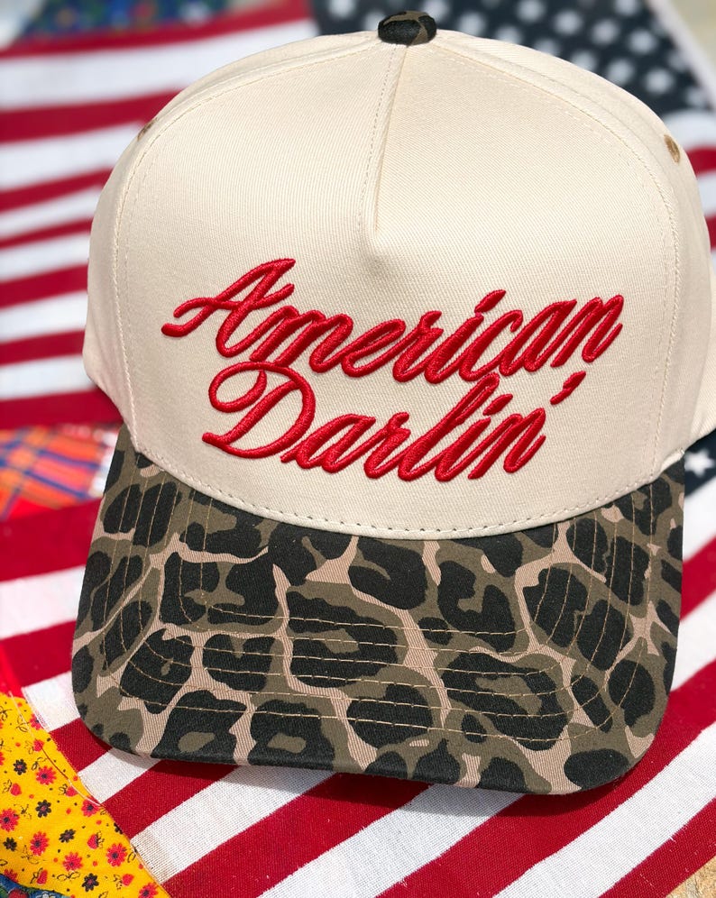 Puede incluir: Una gorra de b&eacute;isbol de color crema con el texto rojo bordado American Darlin' en el panel frontal. La visera de la gorra muestra un patr&oacute;n de estampado de leopardo marr&oacute;n y negro, y un peque&ntilde;o bot&oacute;n a juego se encuentra en la parte superior.