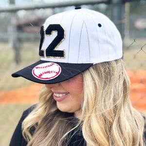 May include: White baseball cap with black brim and the number "12" in black with gold glitter trim. A red and white baseball patch is on the brim. The cap has a pinstripe design.