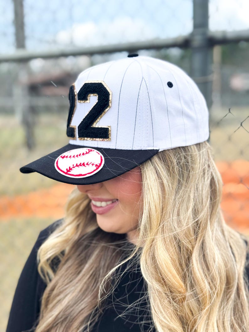May include: White baseball cap with black brim and the number "12" in black with gold glitter trim. A red and white baseball patch is on the brim. The cap has a pinstripe design.