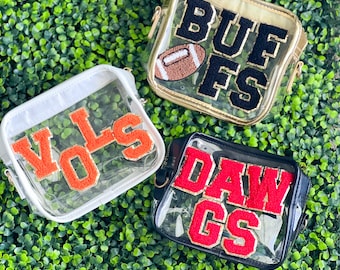 May include: Three clear plastic crossbody bags with different team logos. The first bag is white with an orange "VOLS" logo. The second bag is gold with a black "BUFS" logo and a brown football. The third bag is black with a red "DAWGS" logo.