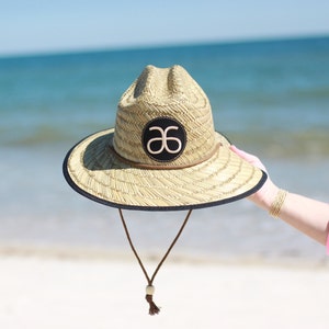 May include: A straw hat with a black band and a brown leather strap. The hat has a logo on the front that says "a6".
