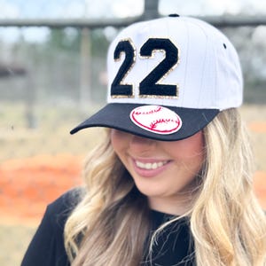 May include: White baseball cap with a black brim, featuring the number "22" in black with gold glitter accents. A baseball graphic is on the front. The cap is worn by a person with blonde hair.
