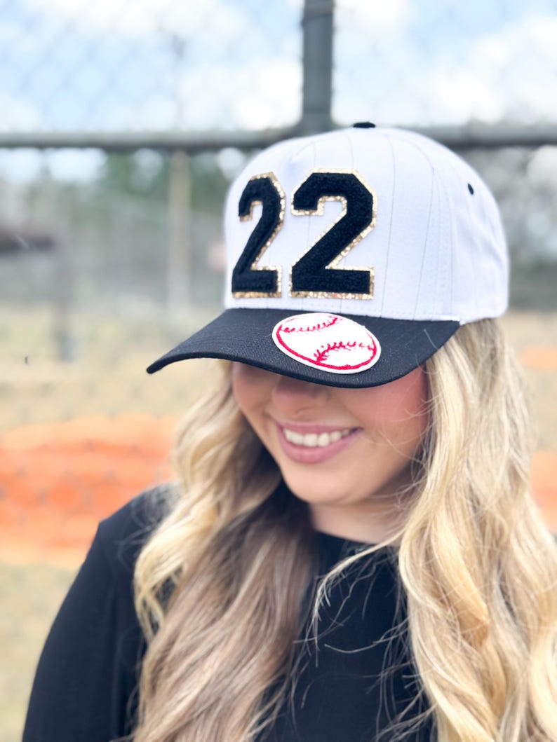 May include: White baseball cap with a black brim, featuring the number "22" in black with gold glitter accents. A baseball graphic is on the front. The cap is worn by a person with blonde hair.