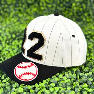 May include: A white baseball cap with black pinstripes and a black brim. The cap features the number "12" in black with gold trim and a baseball patch. A black button sits atop the cap.