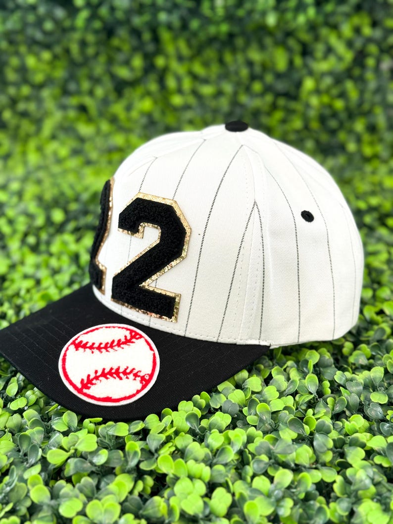May include: A white baseball cap with black pinstripes and a black brim. The cap features the number "12" in black with gold trim and a baseball patch. A black button sits atop the cap.