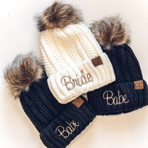 May include: Three knitted beanies with faux fur pom poms. One white beanie says "Bride" and the other two black beanies say "Babe".
