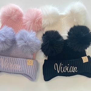 May include: Three knitted hats with faux fur pom-poms. The hats are in pink, light purple, and black. Each hat has the name "Vivian" embroidered on the brim.
