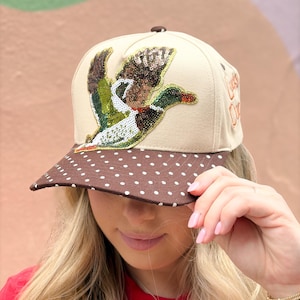 May include: A beige baseball cap featuring a sequined mallard duck appliqué on the front panel. The brim is brown with a white polka dot pattern. The side of the cap displays the embroidered text Lucky Duck in orange thread with a small brown button.