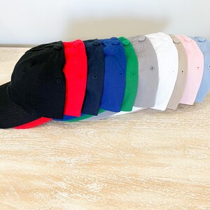 May include: A row of ten blank baseball caps in various colors, including black, red, blue, green, gray, white, beige, pink, and light blue. The caps are arranged in a line, with the black cap at the front and the light blue cap at the back.