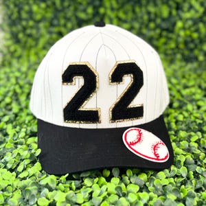 May include: A baseball cap with a black brim and a white crown featuring vertical pinstripes. The number "22" is prominently displayed in black with gold glitter accents. A baseball patch is on the side.