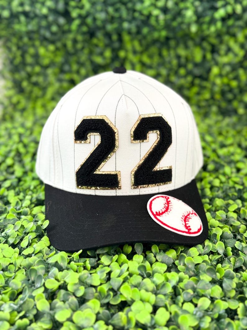 May include: A baseball cap with a black brim and a white crown featuring vertical pinstripes. The number "22" is prominently displayed in black with gold glitter accents. A baseball patch is on the side.