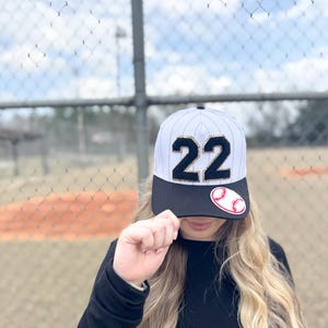 May include: Baseball cap with a black brim and a white and gray striped crown. The number "22" is prominently displayed in black with gold trim. A baseball patch is on the side. The cap is worn by a person.