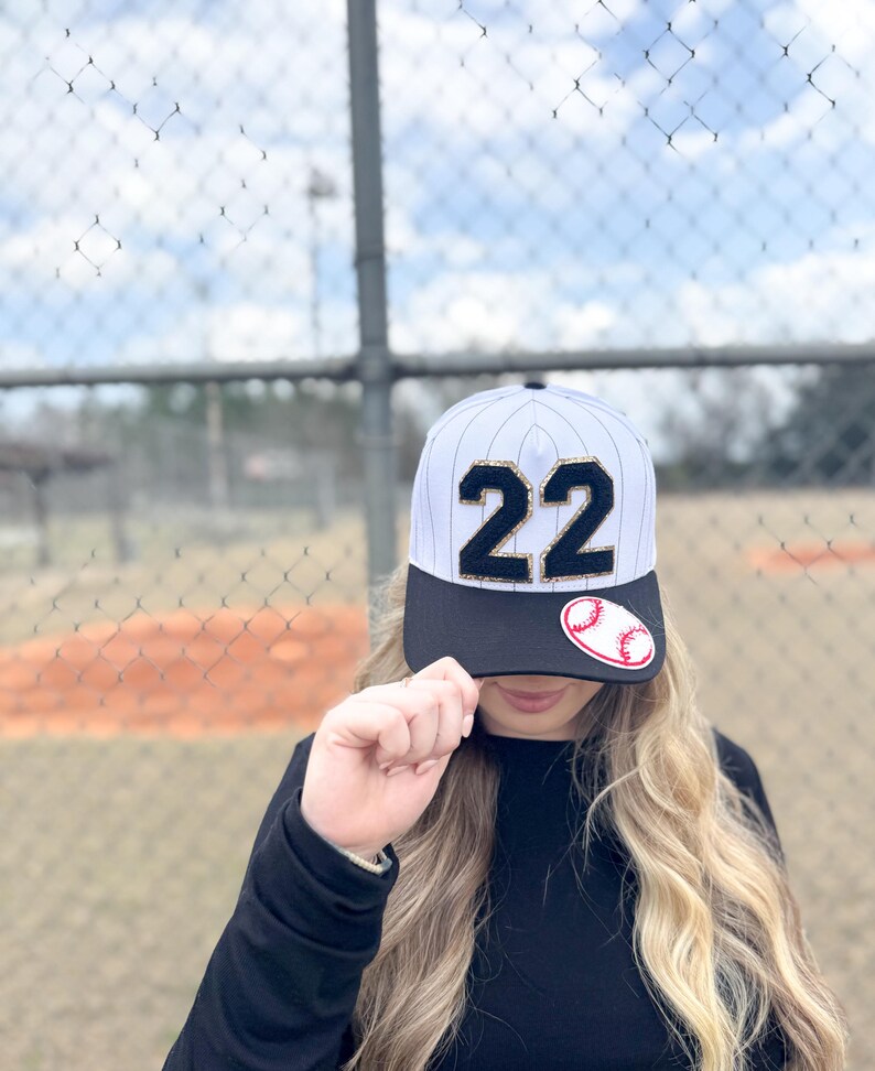 May include: Baseball cap with a black brim and a white and gray striped crown. The number "22" is prominently displayed in black with gold trim. A baseball patch is on the side. The cap is worn by a person.