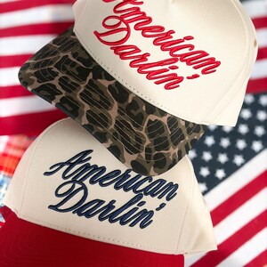 Puede incluir: Dos gorras de b&eacute;isbol apiladas verticalmente. La gorra superior es de color crema con visera de estampado de leopardo y texto bordado en rojo American Darlin'. La gorra inferior es de color crema con visera roja y texto bordado en azul marino American Darlin'.
