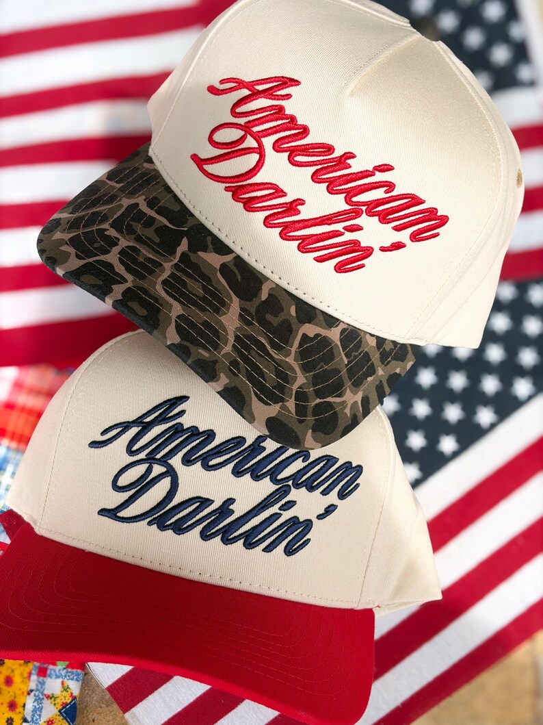 Puede incluir: Dos gorras de b&eacute;isbol apiladas verticalmente. La gorra superior es de color crema con visera de estampado de leopardo y texto bordado en rojo American Darlin'. La gorra inferior es de color crema con visera roja y texto bordado en azul marino American Darlin'.
