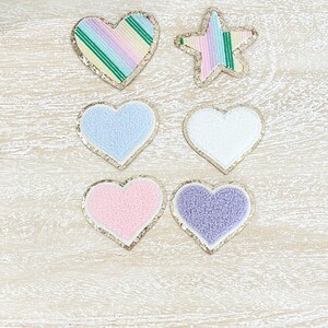 May include: Six heart-shaped patches, four are fuzzy and two are sparkly. The fuzzy patches are in light blue, white, pink, and purple. The sparkly patches are in a rainbow pattern and a green and white striped pattern. There is also a sparkly star-shaped patch in a rainbow pattern.
