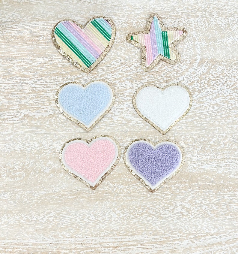 May include: Six heart-shaped patches, four are fuzzy and two are sparkly. The fuzzy patches are in light blue, white, pink, and purple. The sparkly patches are in a rainbow pattern and a green and white striped pattern. There is also a sparkly star-shaped patch in a rainbow pattern.