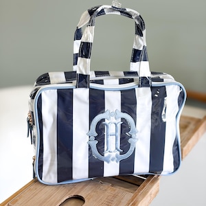 May include: A navy blue and white striped duffel bag with a light blue trim and handles. The bag features a light blue embroidered monogram "H" within a decorative frame. The bag is sitting on a wooden surface.