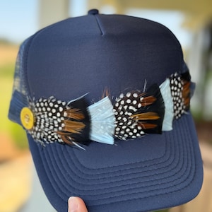 May include: A navy blue trucker hat with a mesh back and a band of feathers across the front. The feathers are brown, white, and black with white spots.