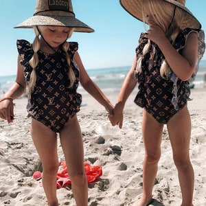 May include: Two young girls wearing matching black and brown swimsuits with a monogram pattern and wide-brimmed straw hats. The hats have a black band with the word "Landrey" written on them. The girls are standing on a sandy beach and holding hands.