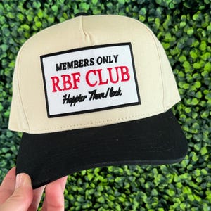May include: A beige and black baseball cap with a black patch that says "Members Only RBF Club Happier Than/Look" in white and red lettering.