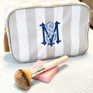 May include: A striped cosmetic bag with a monogram in blue. A makeup brush and a pink compact are in front of the bag. The bag has a gold zipper pull with a tassel.