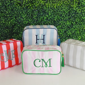 May include: Four striped fabric cosmetic bags with zipper closures and tassels. The bags are in pink and white, red and white, light blue and white, and gray and white. Each bag has a monogram embroidered on the front.