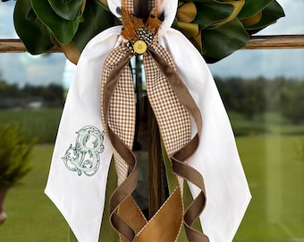 May include: A decorative wreath bow featuring white fabric tails with a green embroidered monogram, brown gingham ribbon, solid brown ribbon, and olive green velvet ribbon. The center is accented with brown feathers and a gold-toned shotgun shell casing.